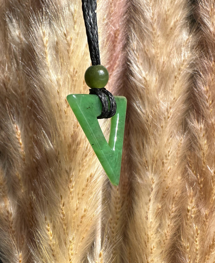 Green jade triangle pendant with a round bead on a black cord against a dried grass backdrop