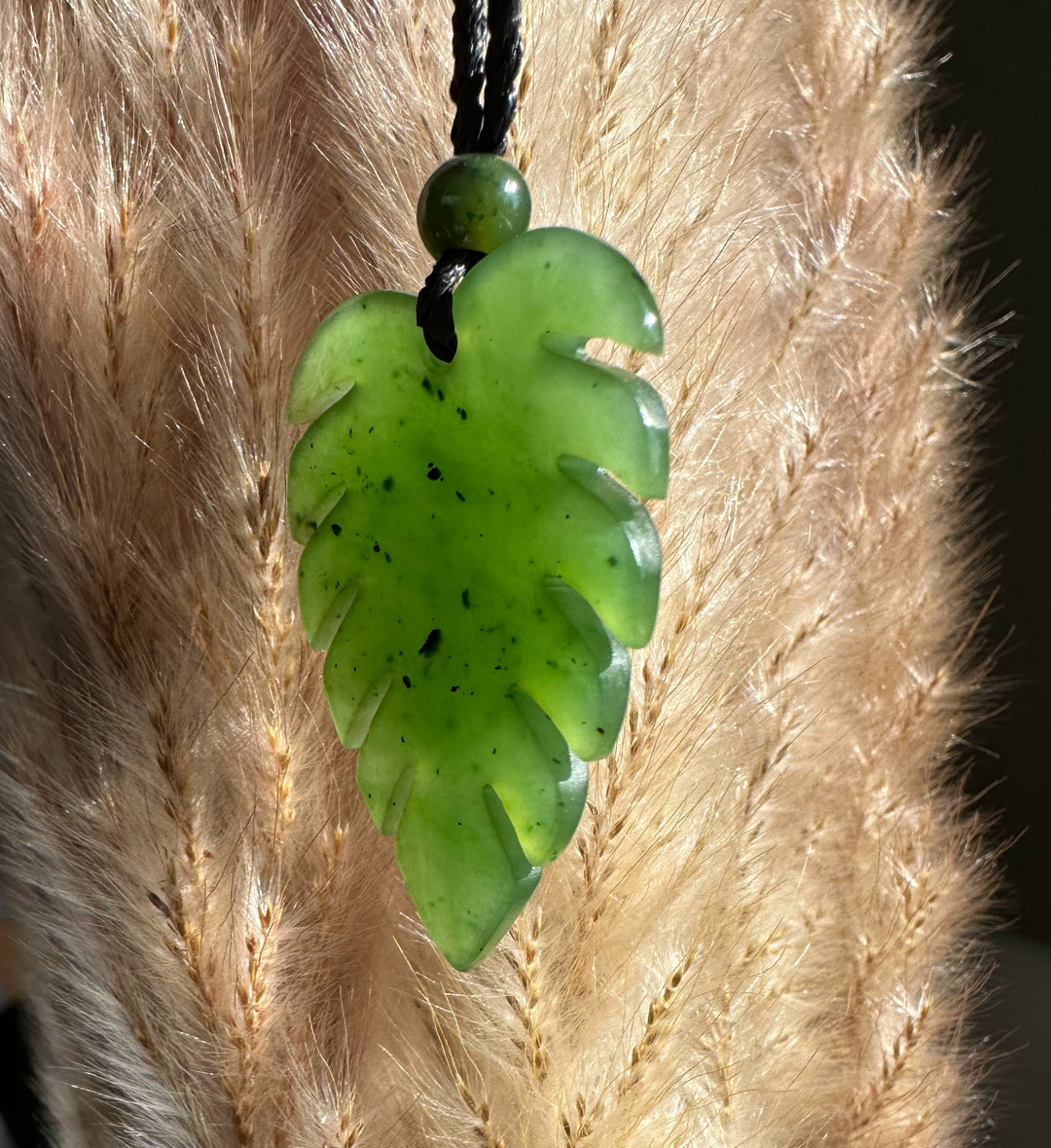 Jade Monstera leaf pendant hanging from a black braided cord with a round bead against a beige background.