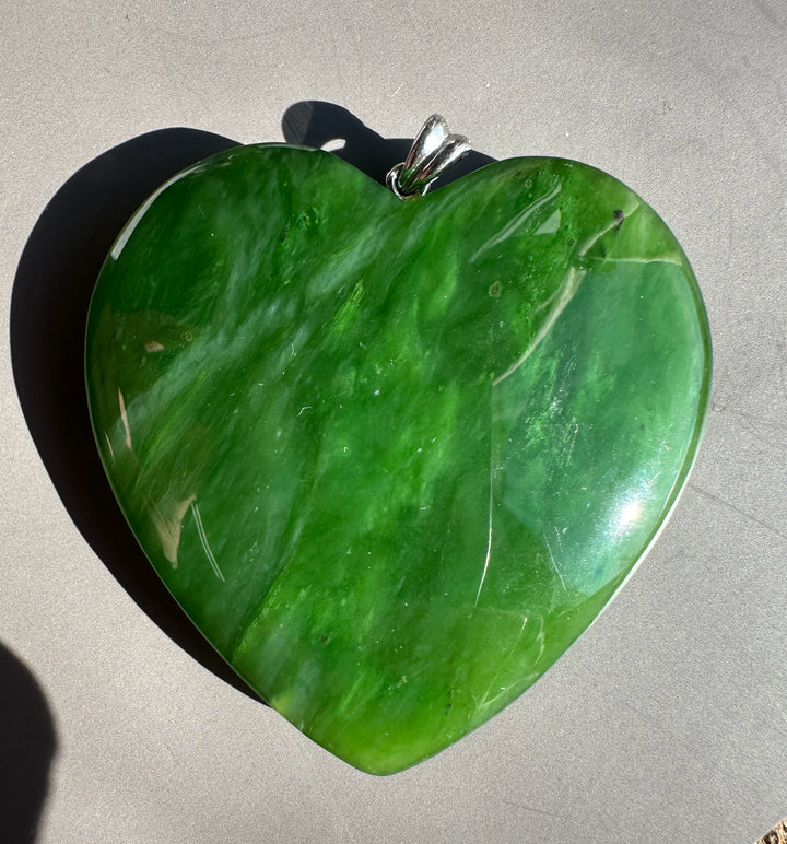 Green polar jade heart pendant with a silver bail resting on a gray surface in sunlight