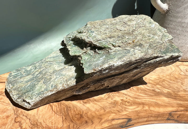 Rough nephrite slab with green jade interior in fractured layers resting on a wooden surface.