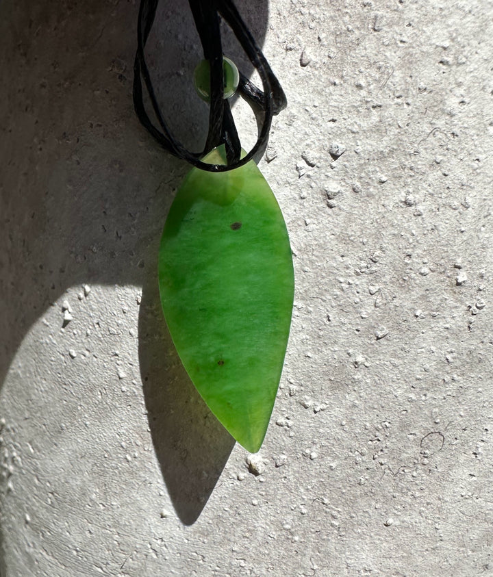 Bright green leaf pendant on a black cord with a small green bead against a textured concrete surface.