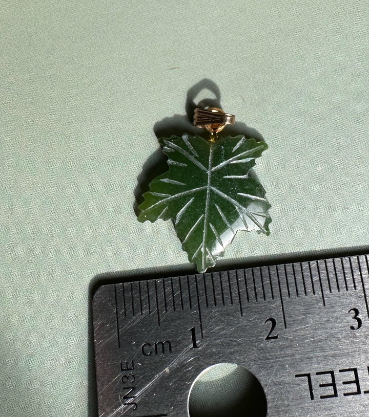 Single dark green maple leaf pendant with gold bail placed on light green fabric beside a ruler