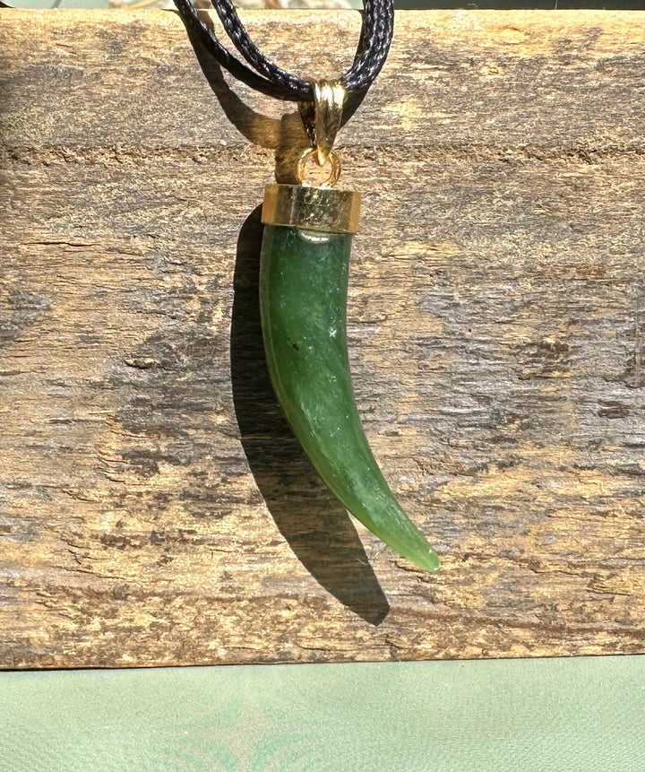 Green jade horn pendant with brass cap on a black cord necklace against a rustic wooden background.