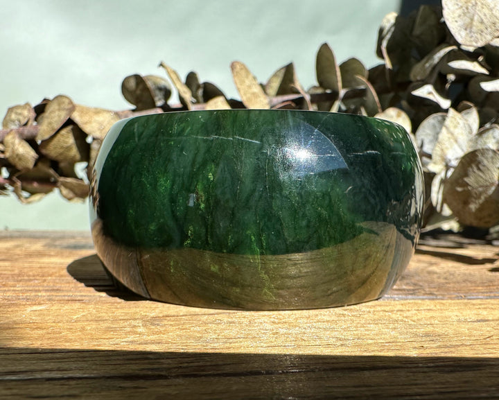 Close up of a glossy dark green jade bangle showing natural banding on a sunlit wooden surface.