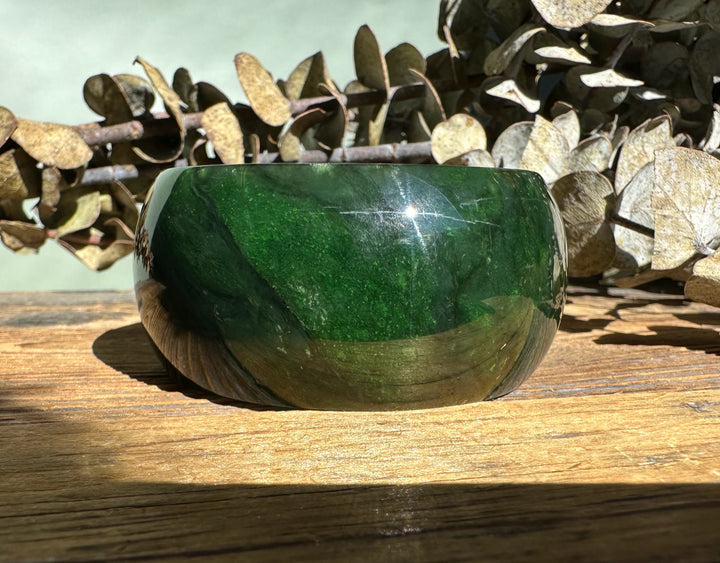 Dark green jade wide bangle with a glossy finish resting on a wooden surface, with dried leaves in the background.