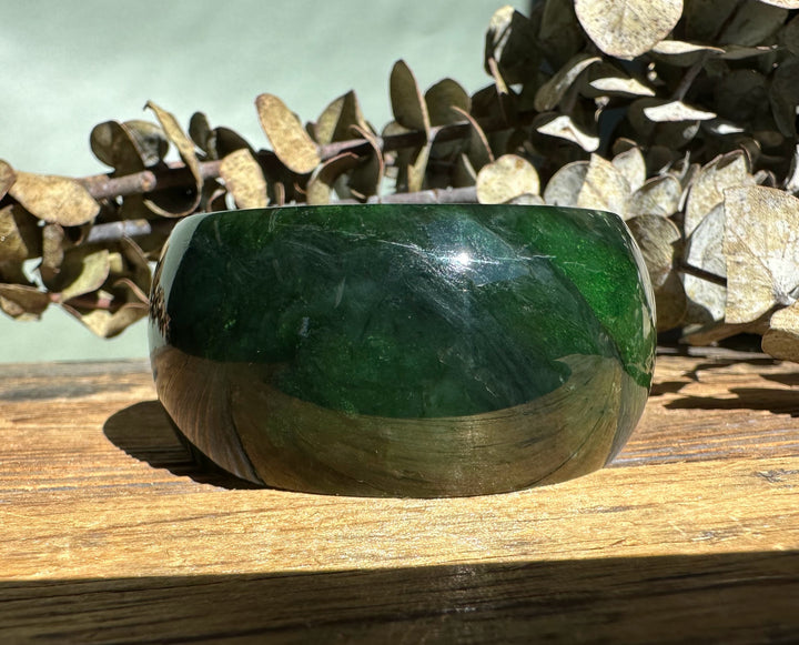 Dark green jade wide bangle with a glossy finish displayed on a sunlit wooden surface.