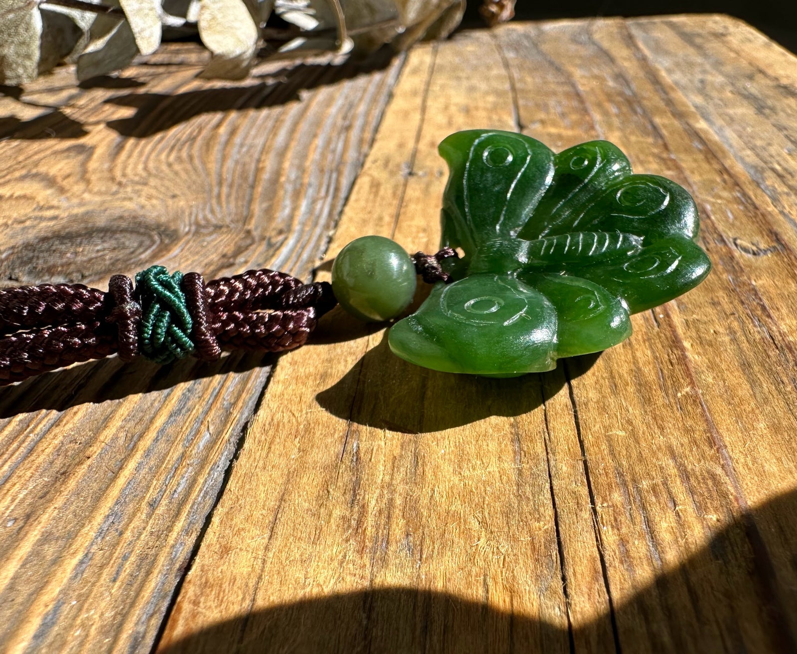 Green jade butterfly pendant on a dark brown braided cord with a round bead, set on a wooden surface