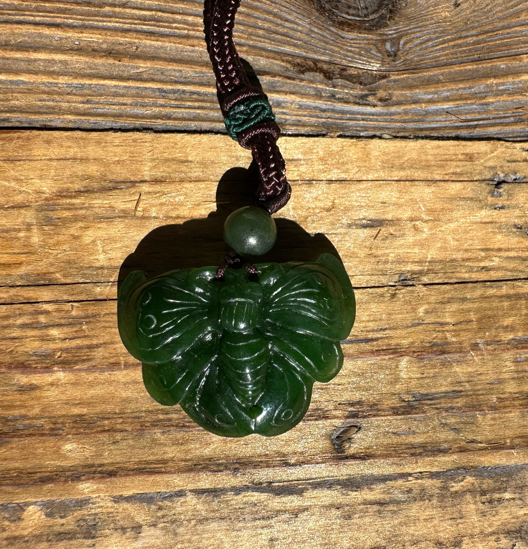 A Grade Ogden Jade Butterfly Pendant in dark green resting on a wooden surface with a brown braided cord and bead