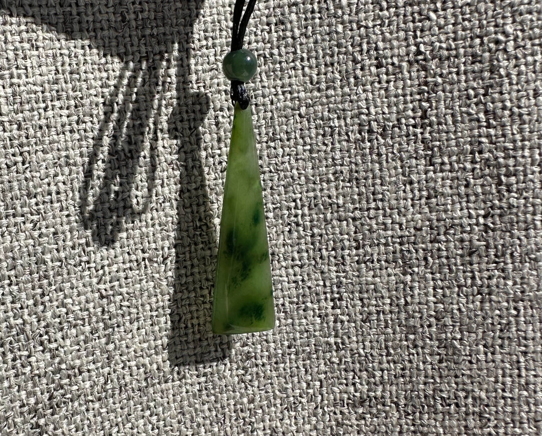 Green jade drop pendant with a round bead on a black cord against gray woven fabric