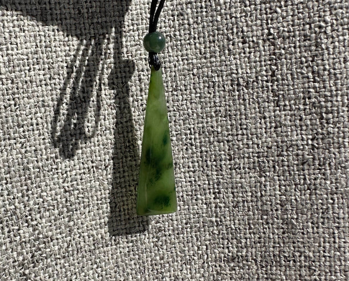 Green jade drop pendant with a round bead on a black cord against gray woven fabric