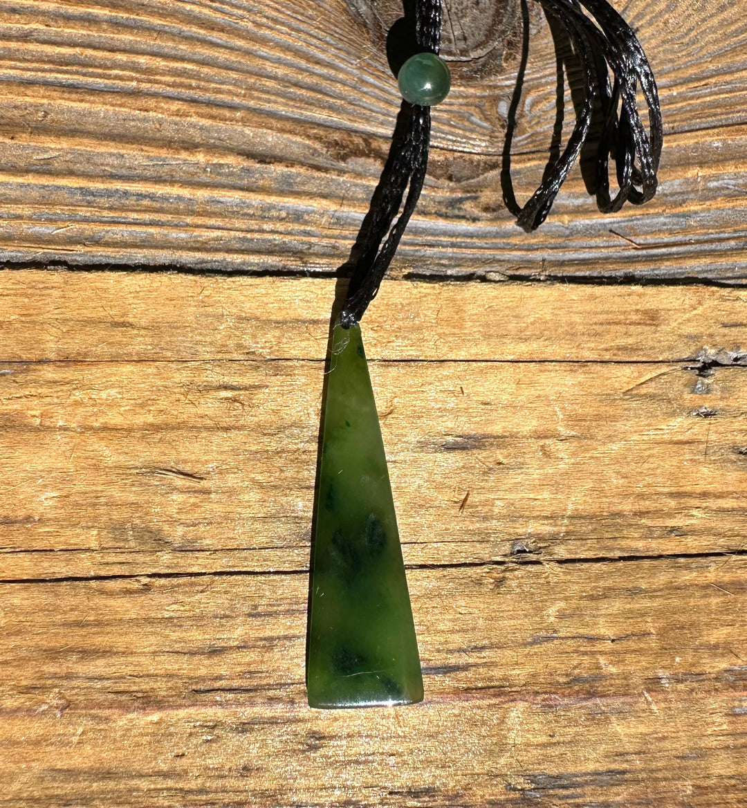 Green jade drop pendant with round bead on black cord shown against rustic wood background