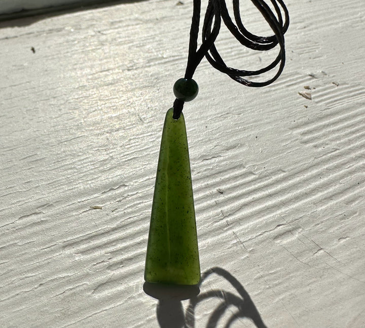 Green jade drop pendant with round bead on black cord resting on pale wooden surface