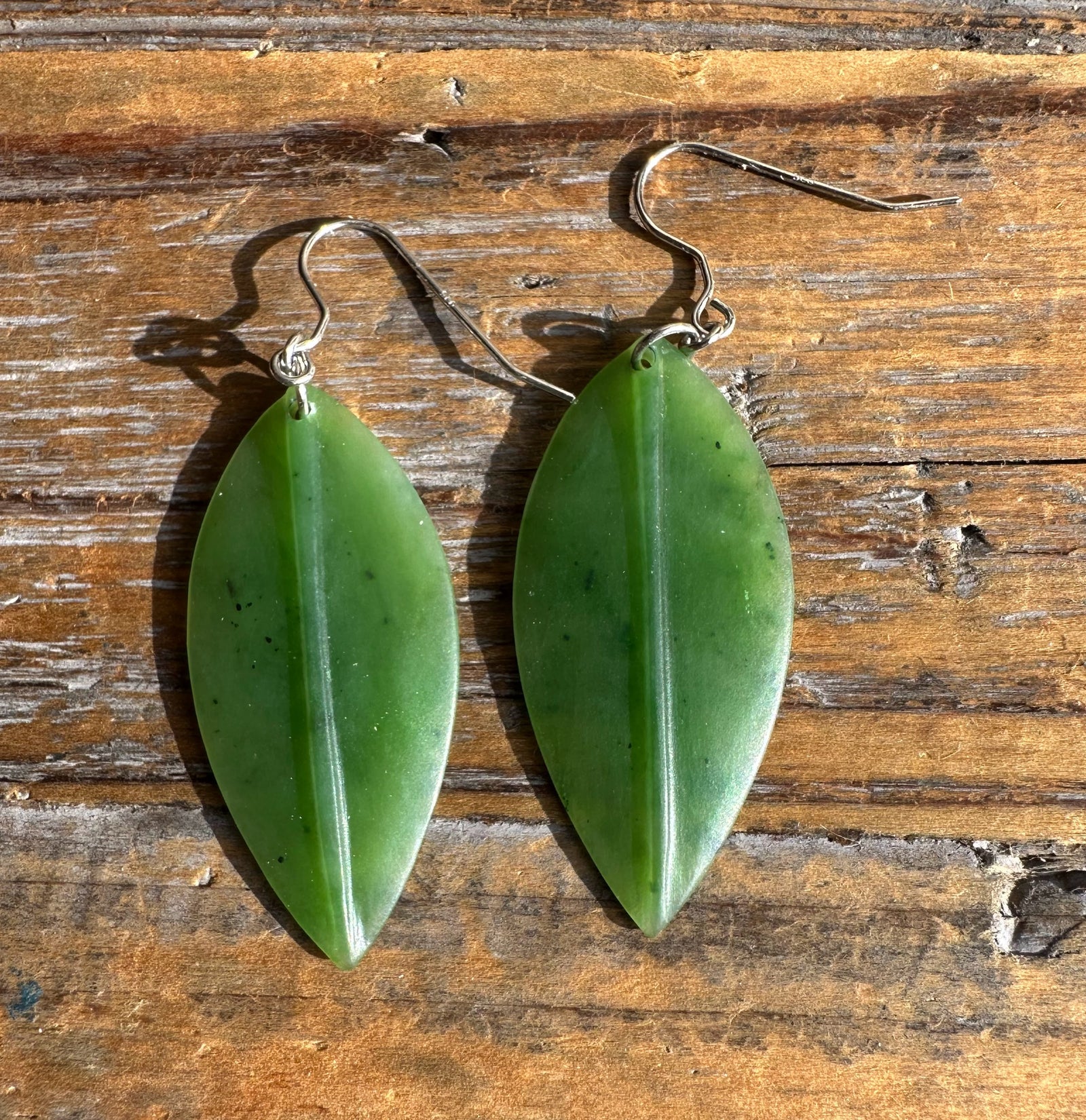 Two jade leaf earrings with green leaves and metal hooks laid on a wooden surface.