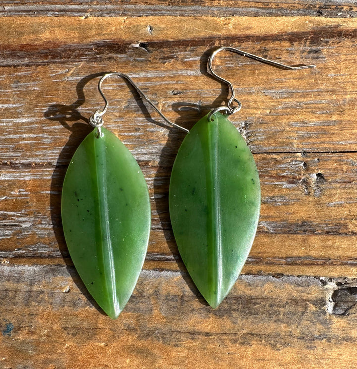 Two jade leaf earrings with green leaves and metal hooks laid on a wooden surface.