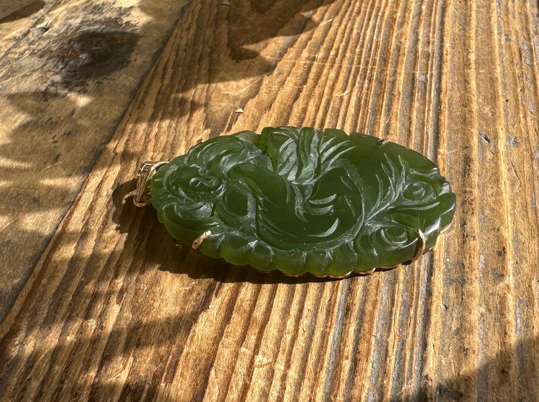 Green jade pendant carved with roses and flowing leaves on a wood surface.