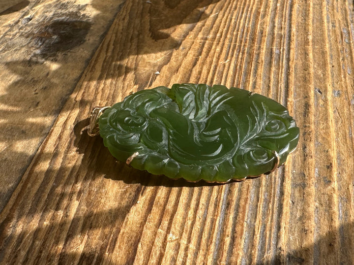 Green jade pendant carved with roses and flowing leaves on a wood surface.