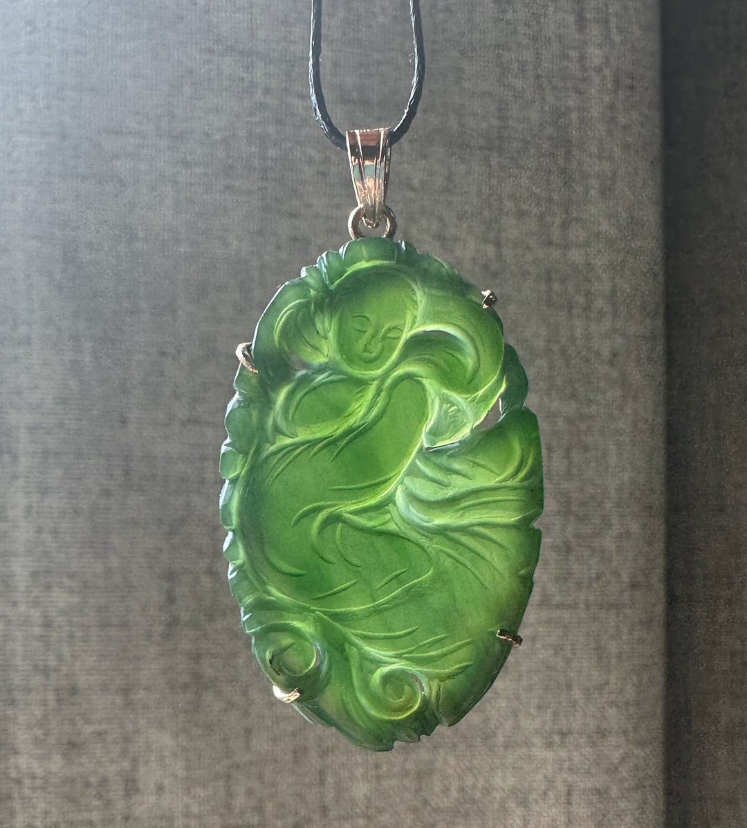 Bright green jade Guanyin pendant with a seated figure carved in relief in a decorative metal frame on a black cord.