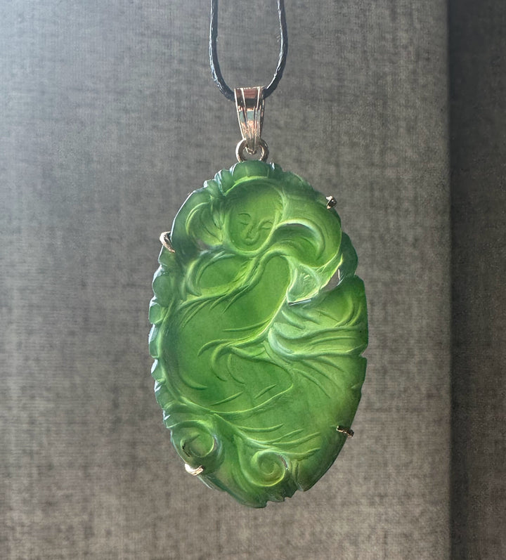 Bright green jade Guanyin pendant with a seated figure carved in relief in a decorative metal frame on a black cord.