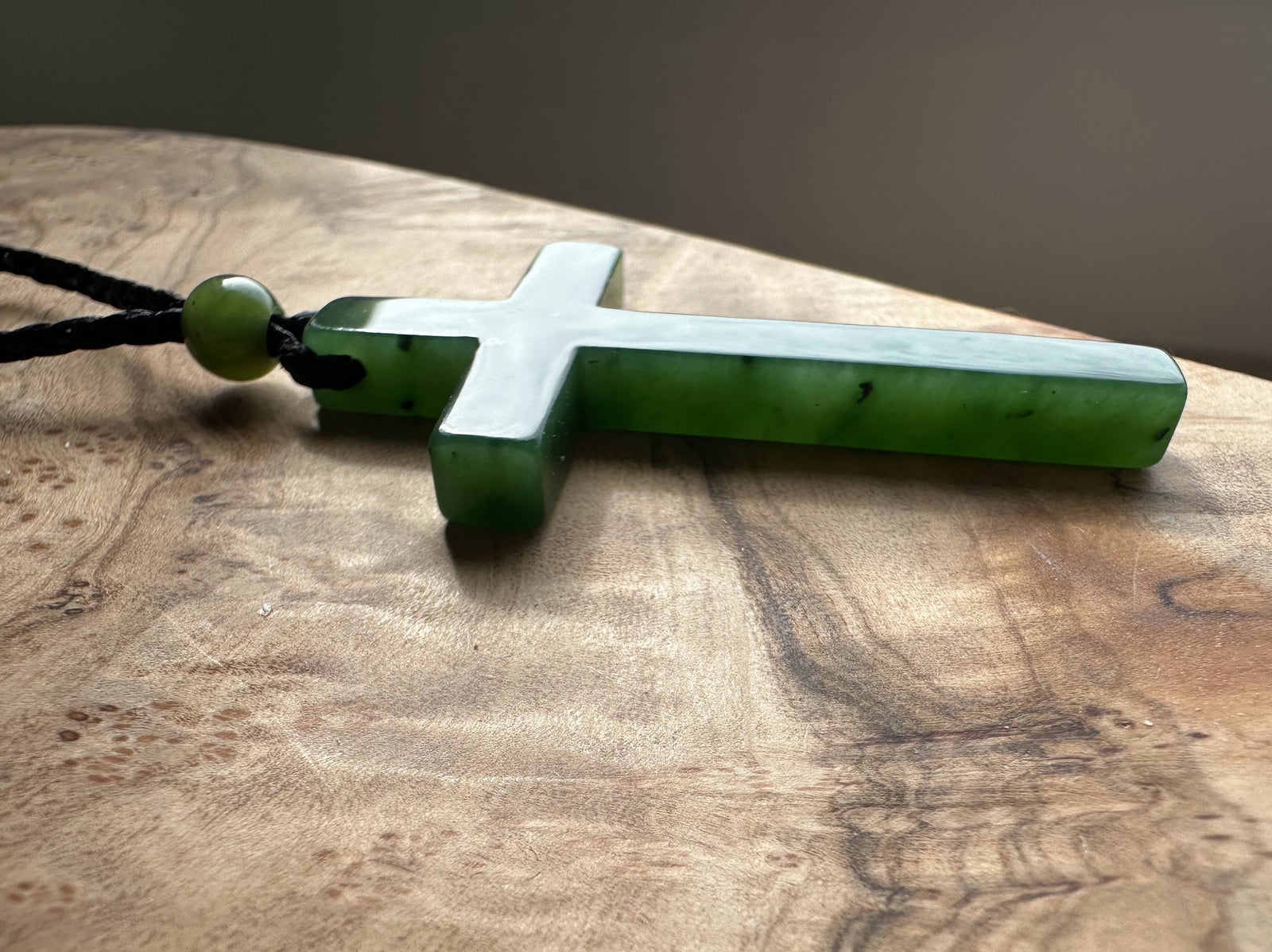 Green jade cross pendant on a black braided cord resting on a wooden surface with a green bead near the loop.