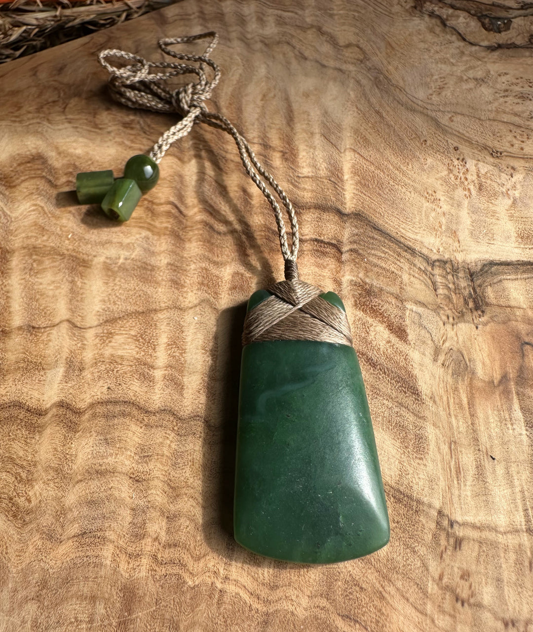 Dark green jade adze pendant with brown cord wrapping and bead accents against a wooden surface
