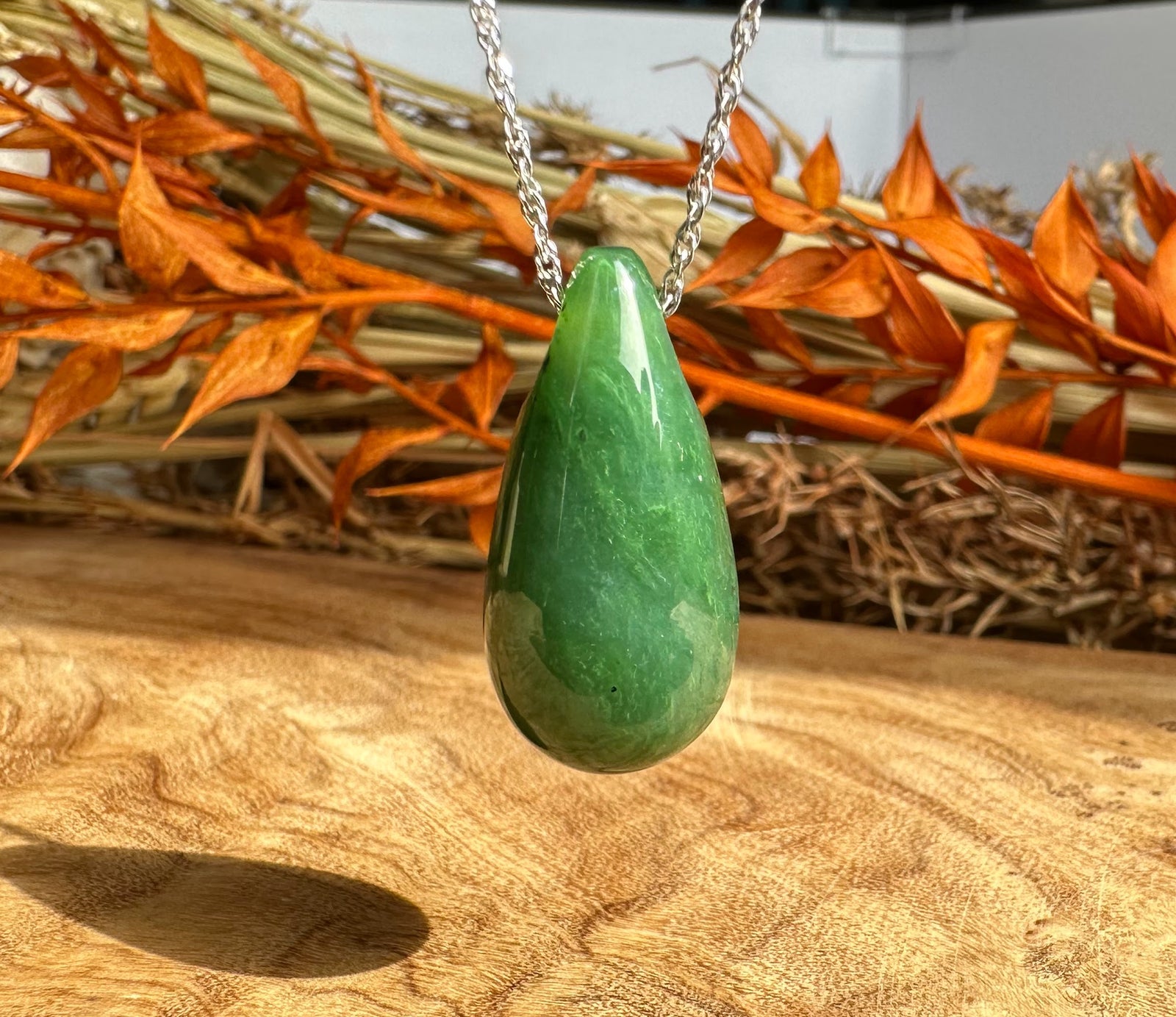 Green jade teardrop pendant on a silver chain against a wooden surface with dried orange leaves
