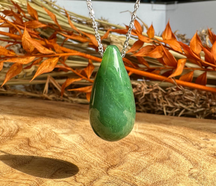 Green jade teardrop pendant on a silver chain against a wooden surface with dried orange leaves
