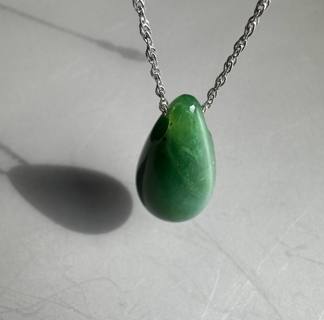 Close up of green jade teardrop pendant on a silver chain against a pale gray backdrop.
