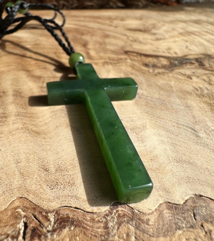 Green jade cross pendant on a black cord angled on a wood surface with a round green bead near the top.
