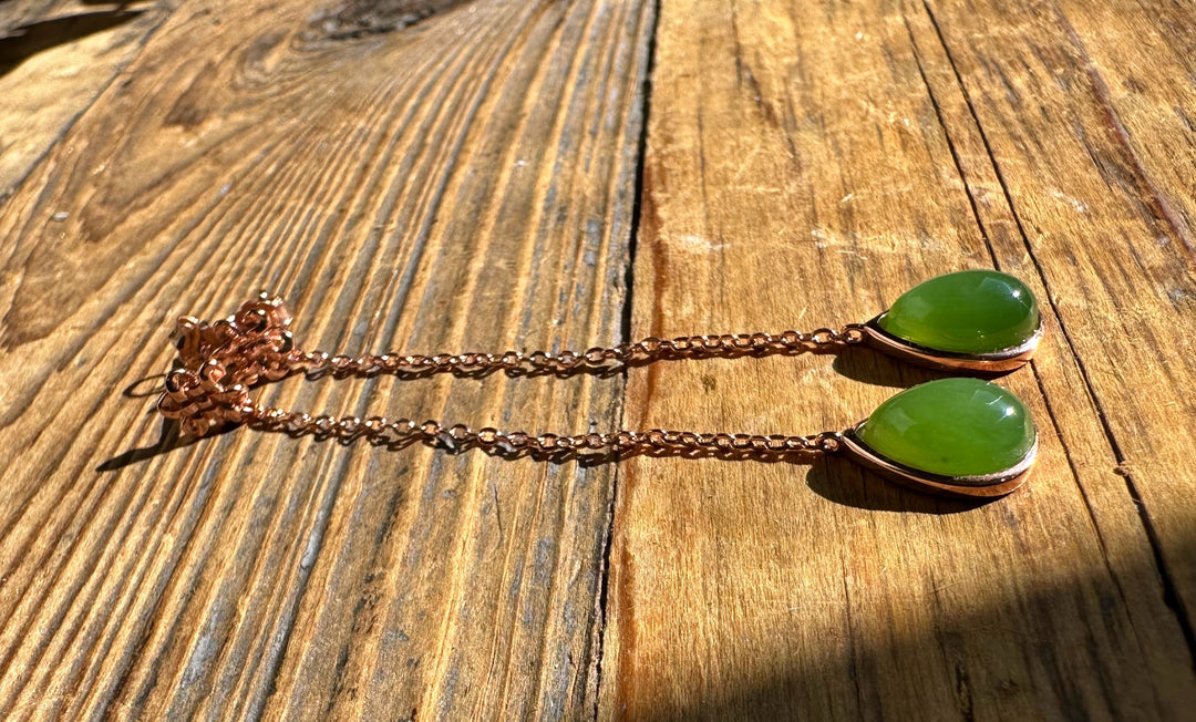 Green jade teardrop pendant earrings with copper bezels on slim copper chains laid on a wooden surface.