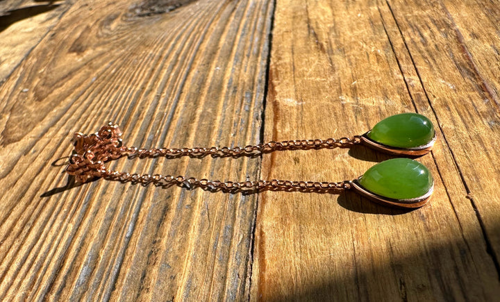 Green jade teardrop pendant earrings with copper bezels on slim copper chains laid on a wooden surface.
