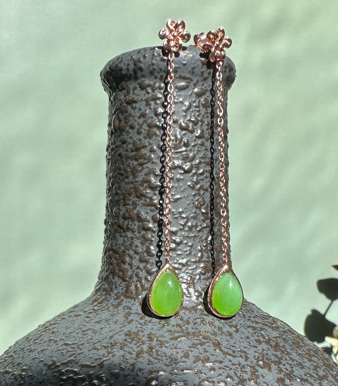 Rose gold chain drop earrings with bright green jade teardrops and flower stud tops, displayed on a textured pedestal.