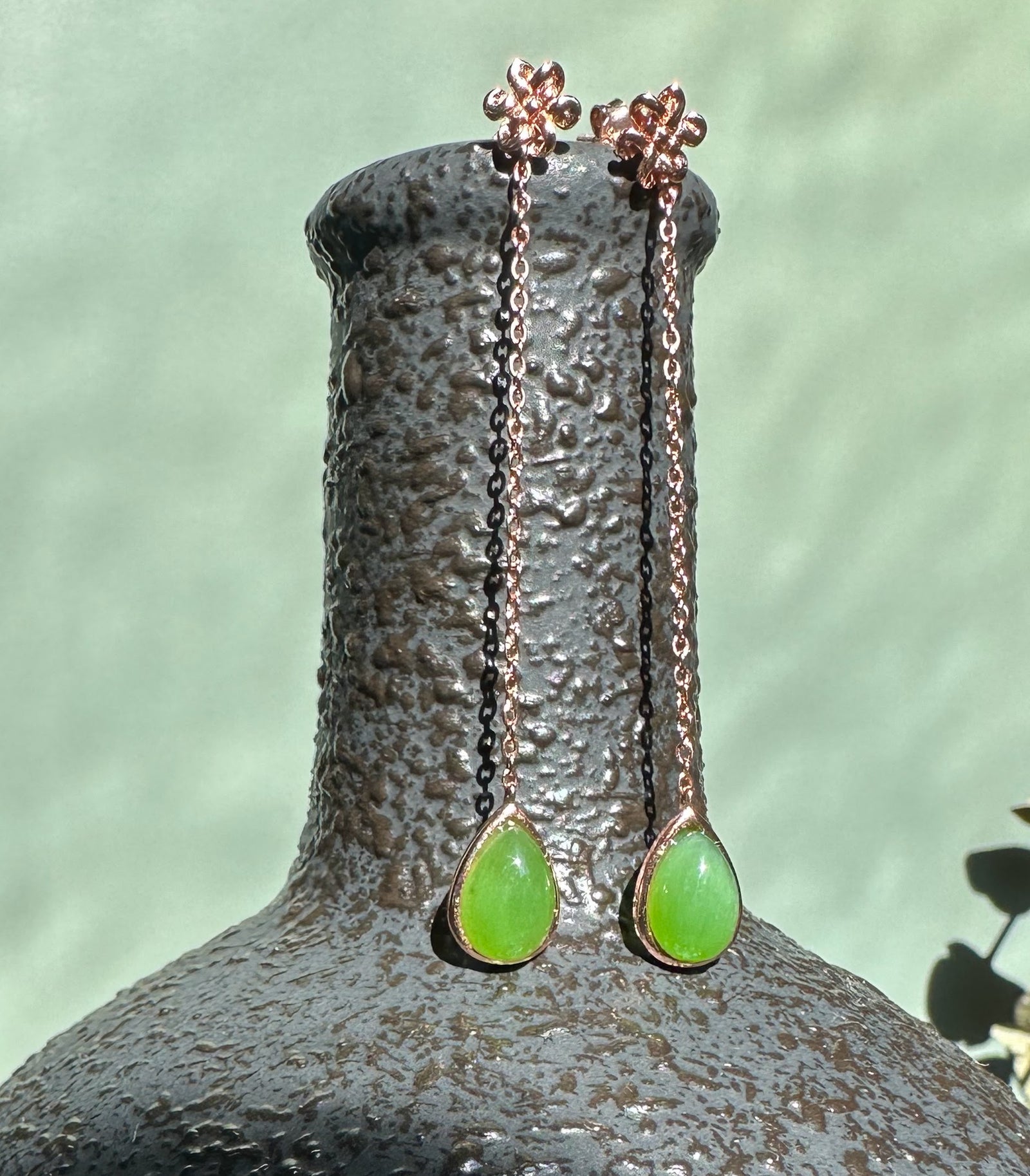 Rose gold chain drop earrings with bright green jade teardrops and flower stud tops, displayed on a textured pedestal.
