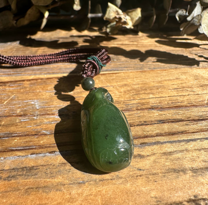 Dark green jade bird pendant with a round bead on a brown braided cord resting on a wooden surface