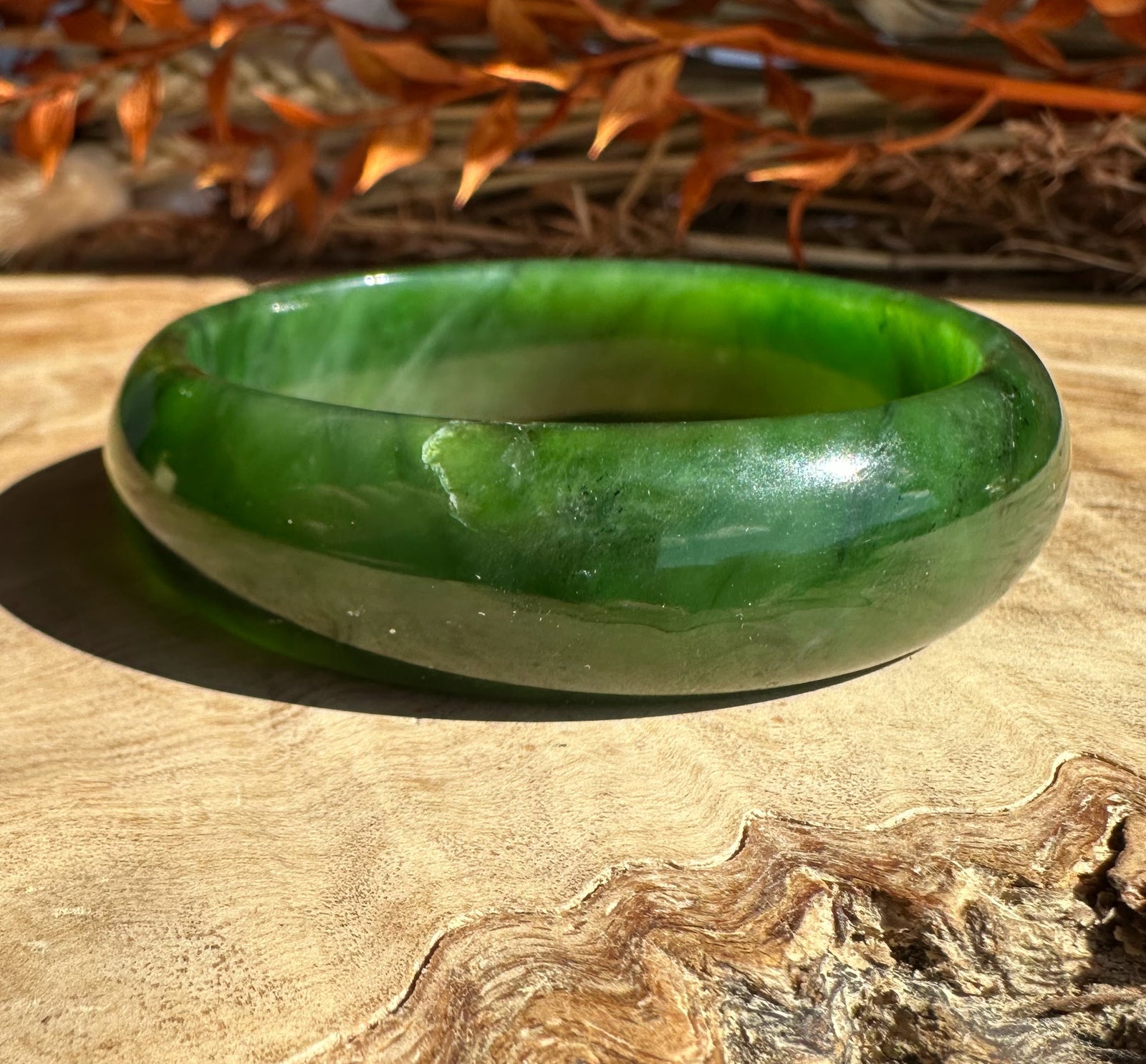 Green jade bangle with a lighter band around it set on wood with dried orange leaves in the background.