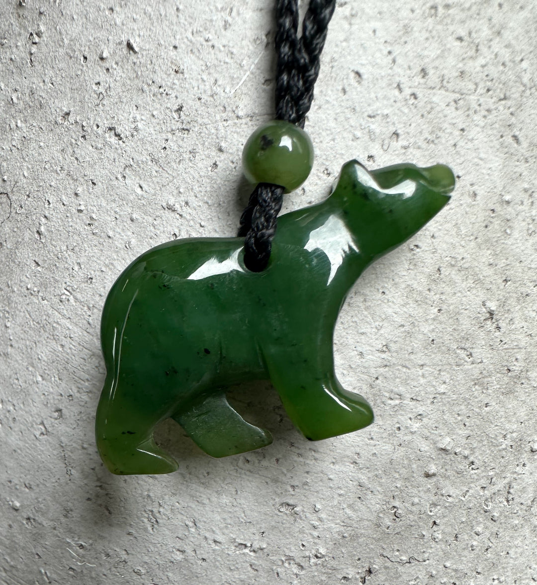 Close up of green jade bear pendant with green bead on a black cord against a light textured surface