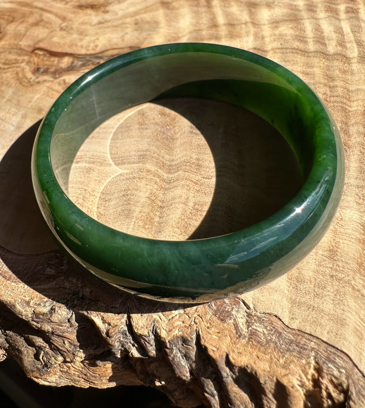 Canadian Jade Bangle 54 x 17mm in dark green rests on a textured wooden surface under sunlight.