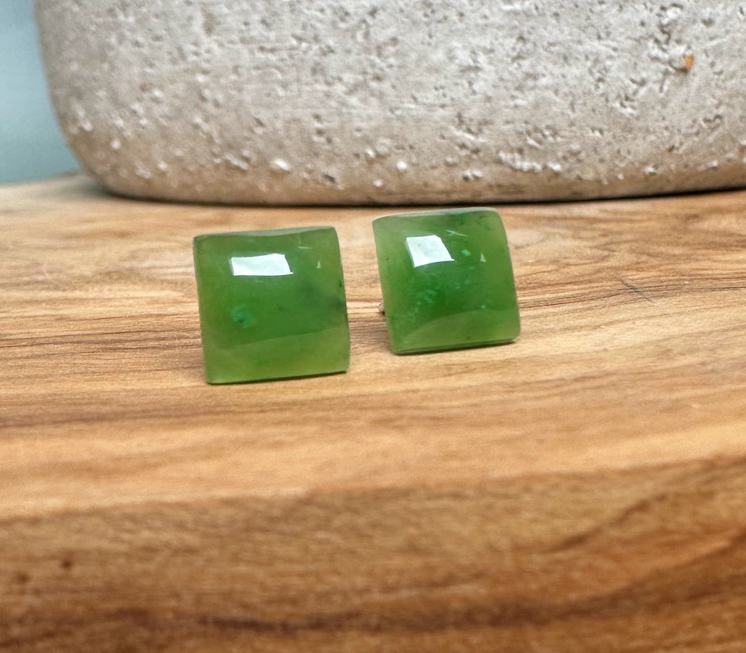 Two green jade square earrings lie on a rustic wooden surface.