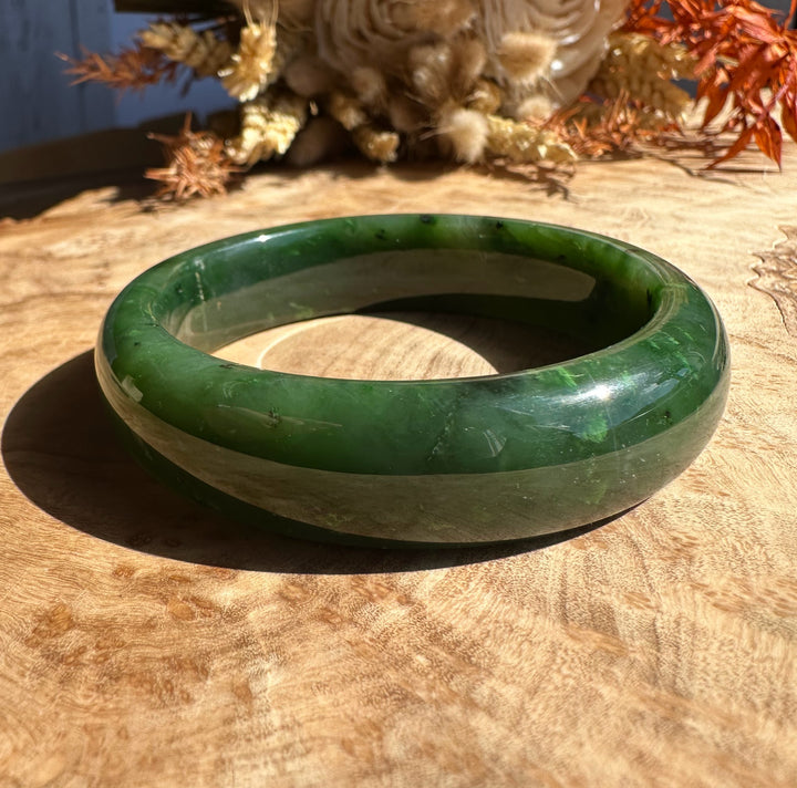 Canadian Jade Bangle 56 x 15.5mm in polished green jade on a wooden surface with dried flowers in the background