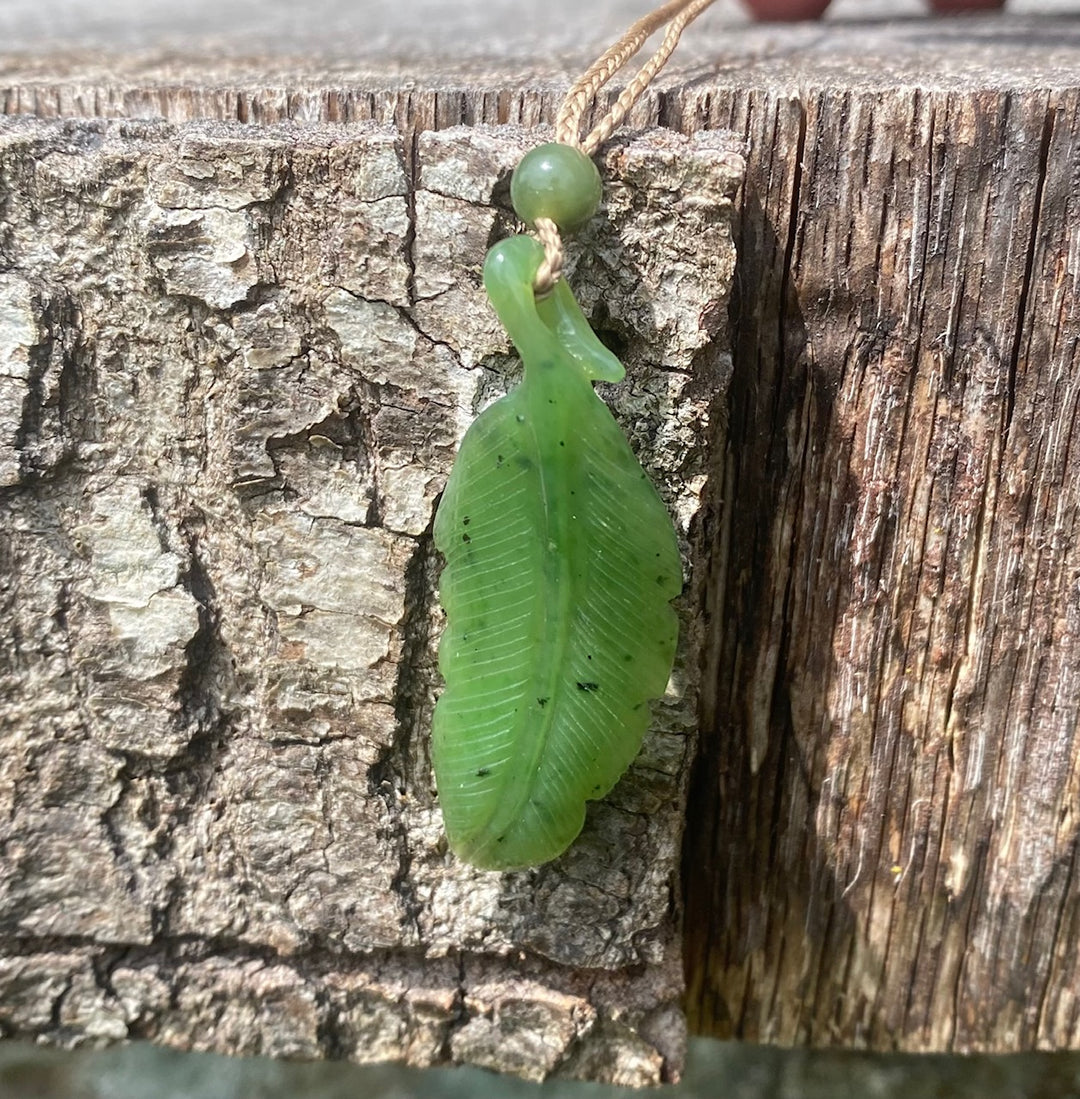 Green leaf shaped jade pendant with veined texture hanging on a beige cord against weathered wood