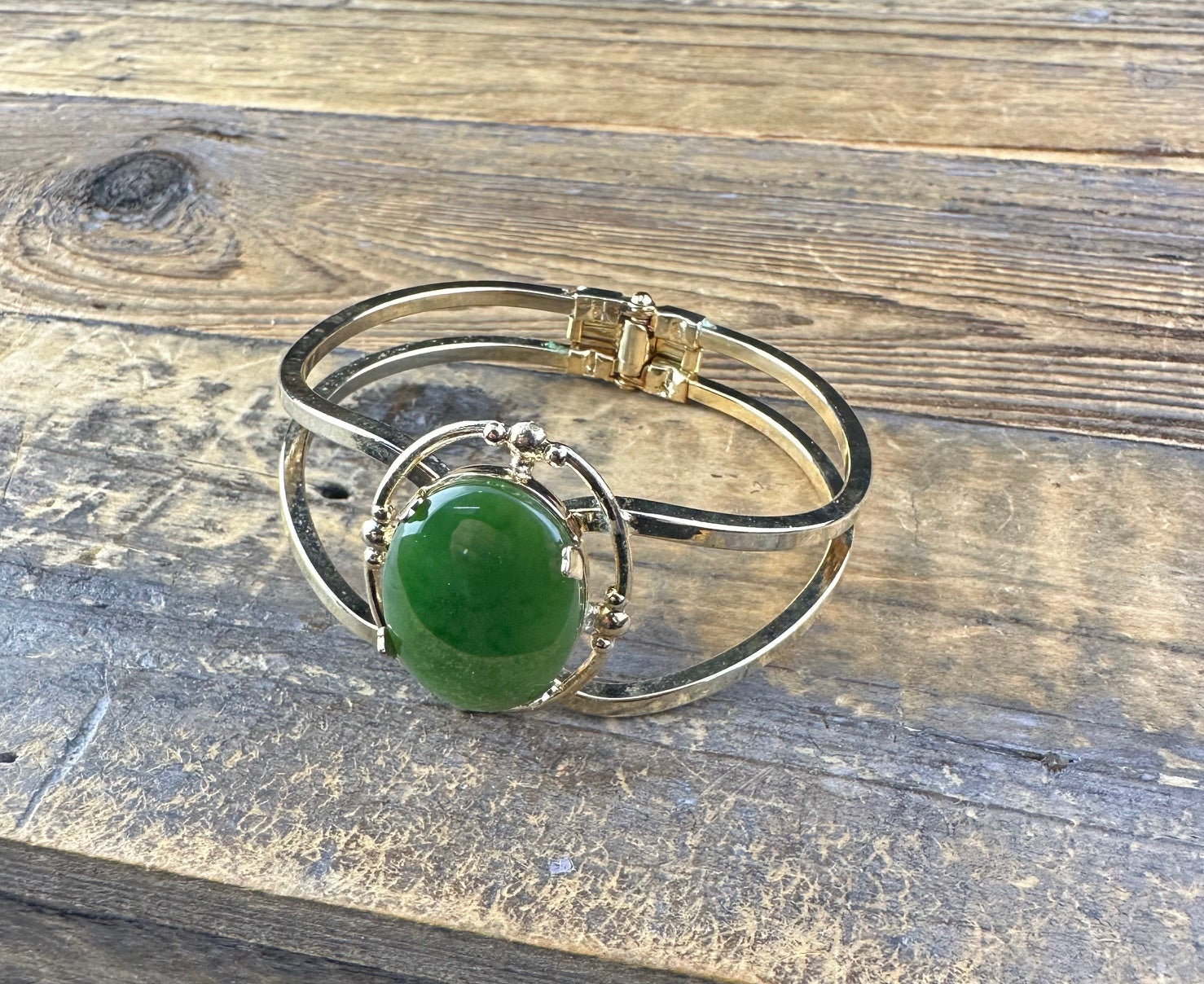 Two-tone vintage bracelet with a large green jade cabochon in a beaded bezel on an open cuff resting on wood