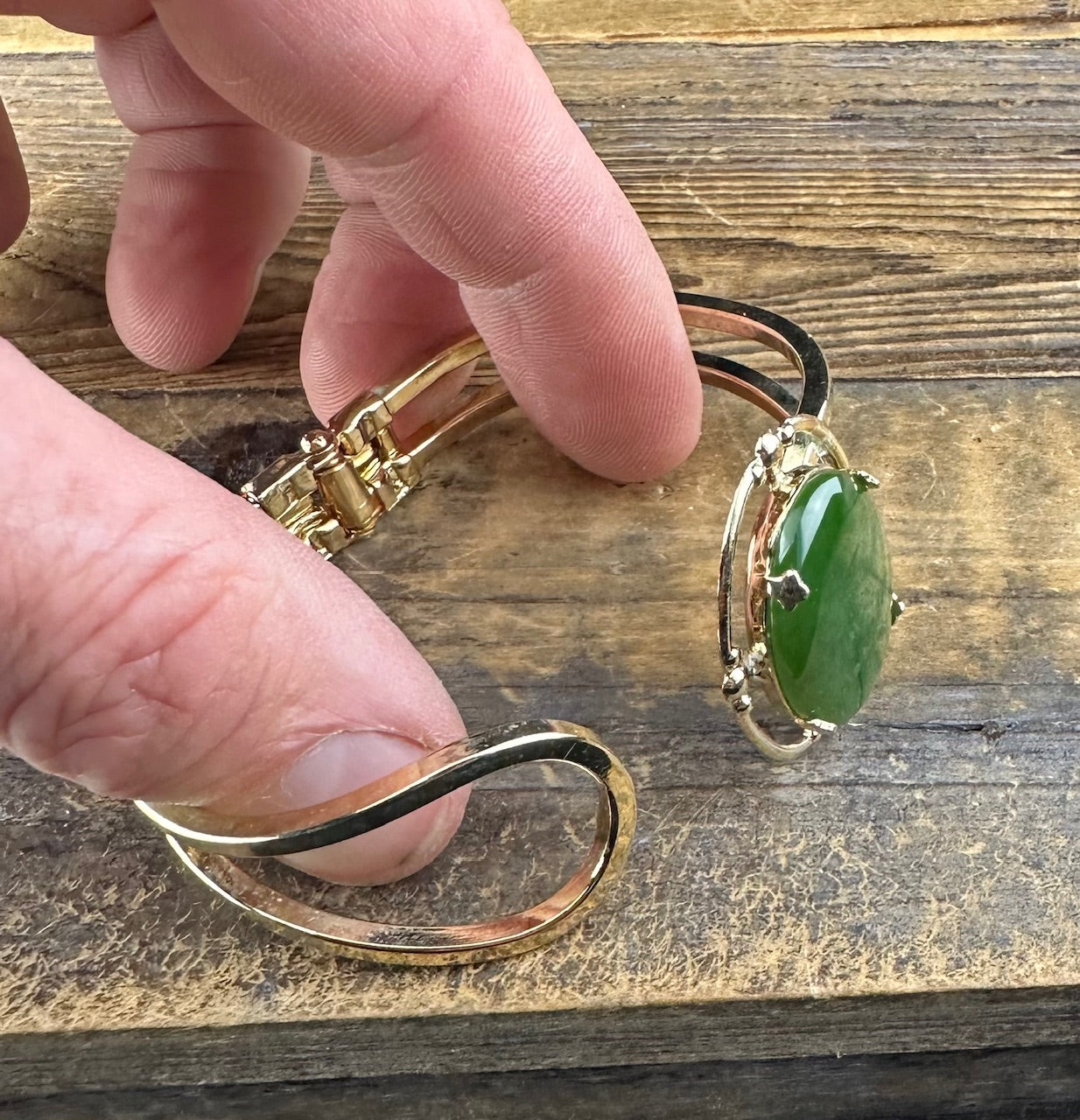 Close-up of a gold-tone vintage jade bracelet showing a large green jade centerpiece held by fingers against a wooden surface