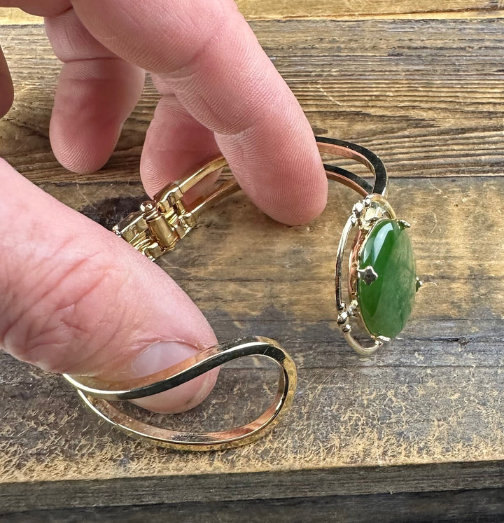 Close-up of a gold-tone vintage jade bracelet showing a large green jade centerpiece held by fingers against a wooden surface