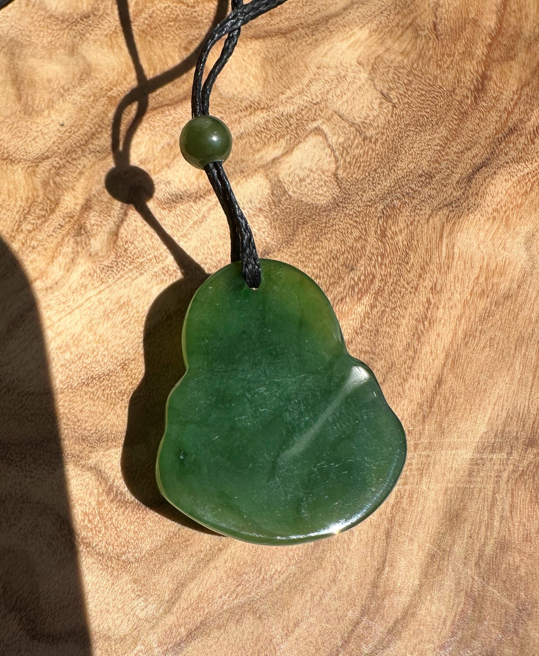 Green jade Laughing Buddha pendant on a black cord with a bead against a wood background