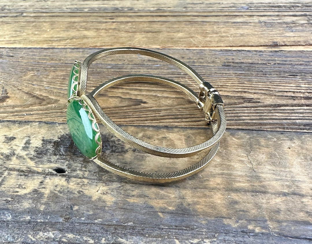 Vintage jade bracelet with a large green oval cabochon set in a gold decorative bezel on a wooden surface.