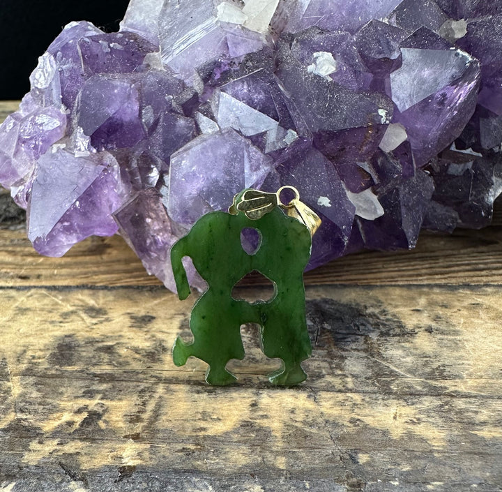 Green jade Young Love Pendant carved into an abstract silhouette with holes, gold bail, in front of a purple amethyst cluster.