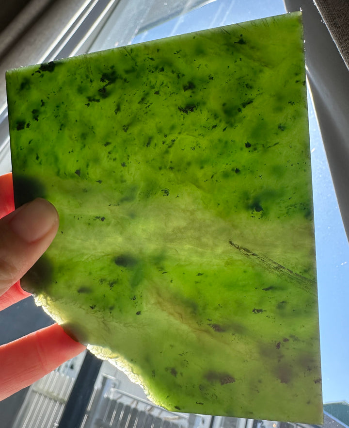 Bright lime green A Grade Canadian Jade Slab 205g with dark flecks held by fingers near a window.