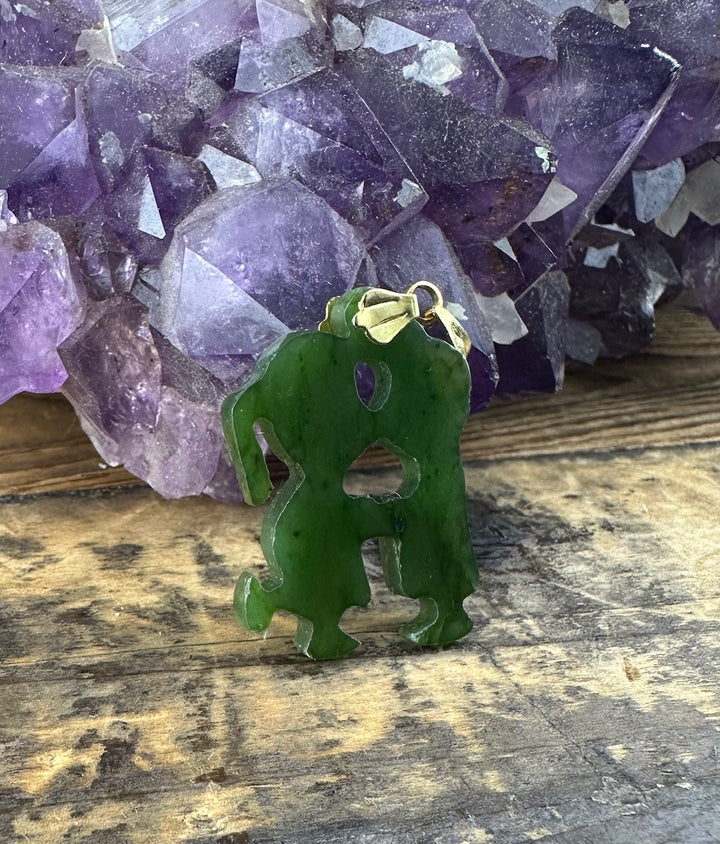 Vintage Jade Young Love Pendant in green jade with gold bail rests on rustic wood with purple amethyst behind.