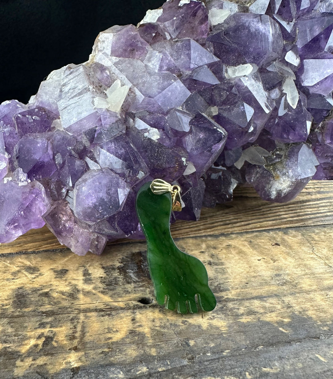 Green jade foot pendant with gold bail displayed on rustic wood near purple amethyst cluster