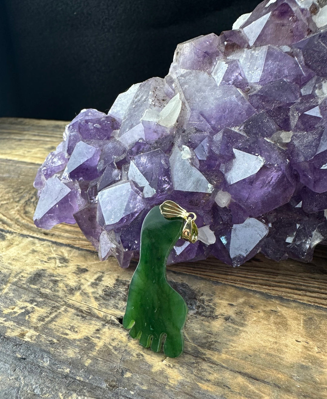 Vintage jade Big Foot pendant in green with a gold bail held in front of a purple amethyst cluster
