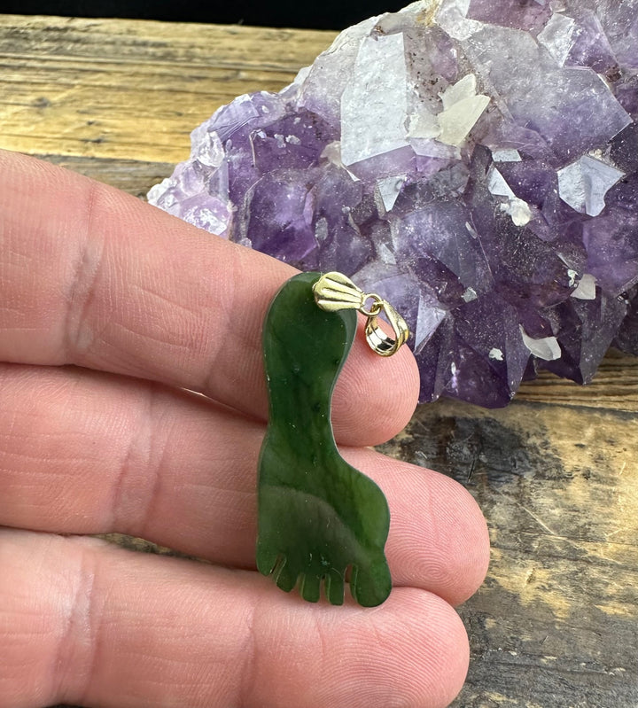 Green jade Big Foot pendant with a gold bail held between fingers beside an amethyst cluster on a wooden surface